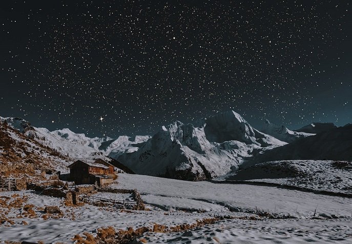 Snow‑covered mountains and a small wooden hut under a star‑filled night sky in Hanle, Ladakh, India
