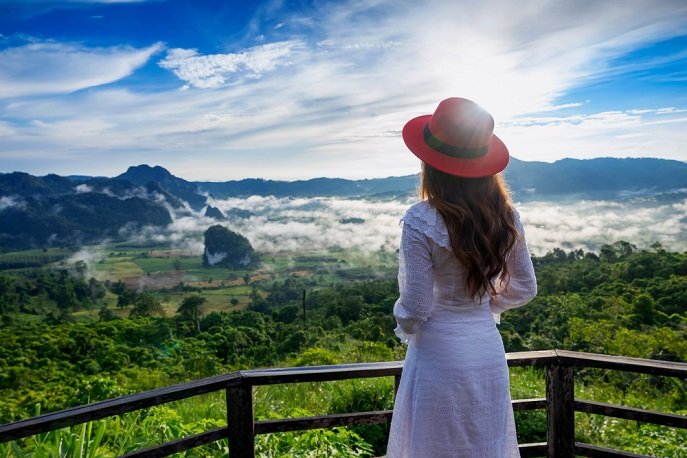 Woman in white dress standing at a distance, watching misty mountains and clouds.