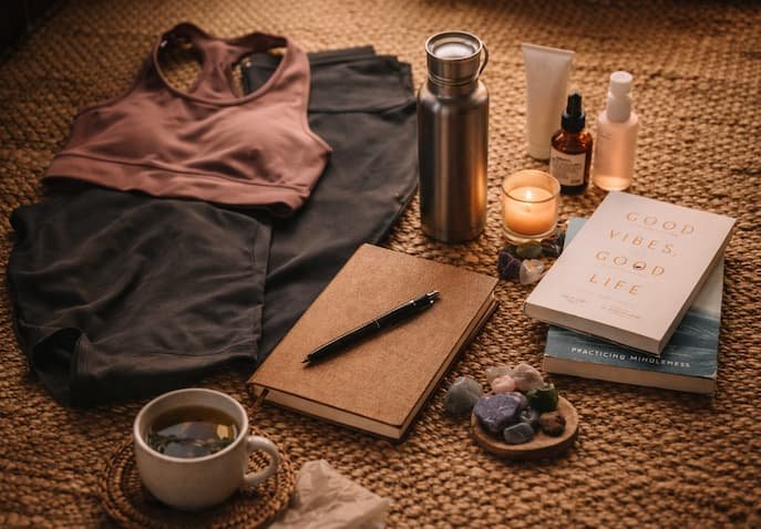 Packing essentials for women are kept like a journal, a book, skincare items, herbal tea, & activewear.