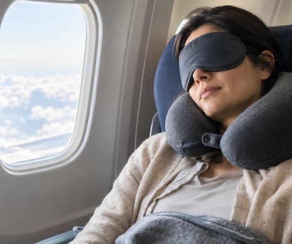 A woman sleeping peacefully with an eye mask and neck pillow on flight. She is through with tips that help explain how to sleep better on long-haul flights.